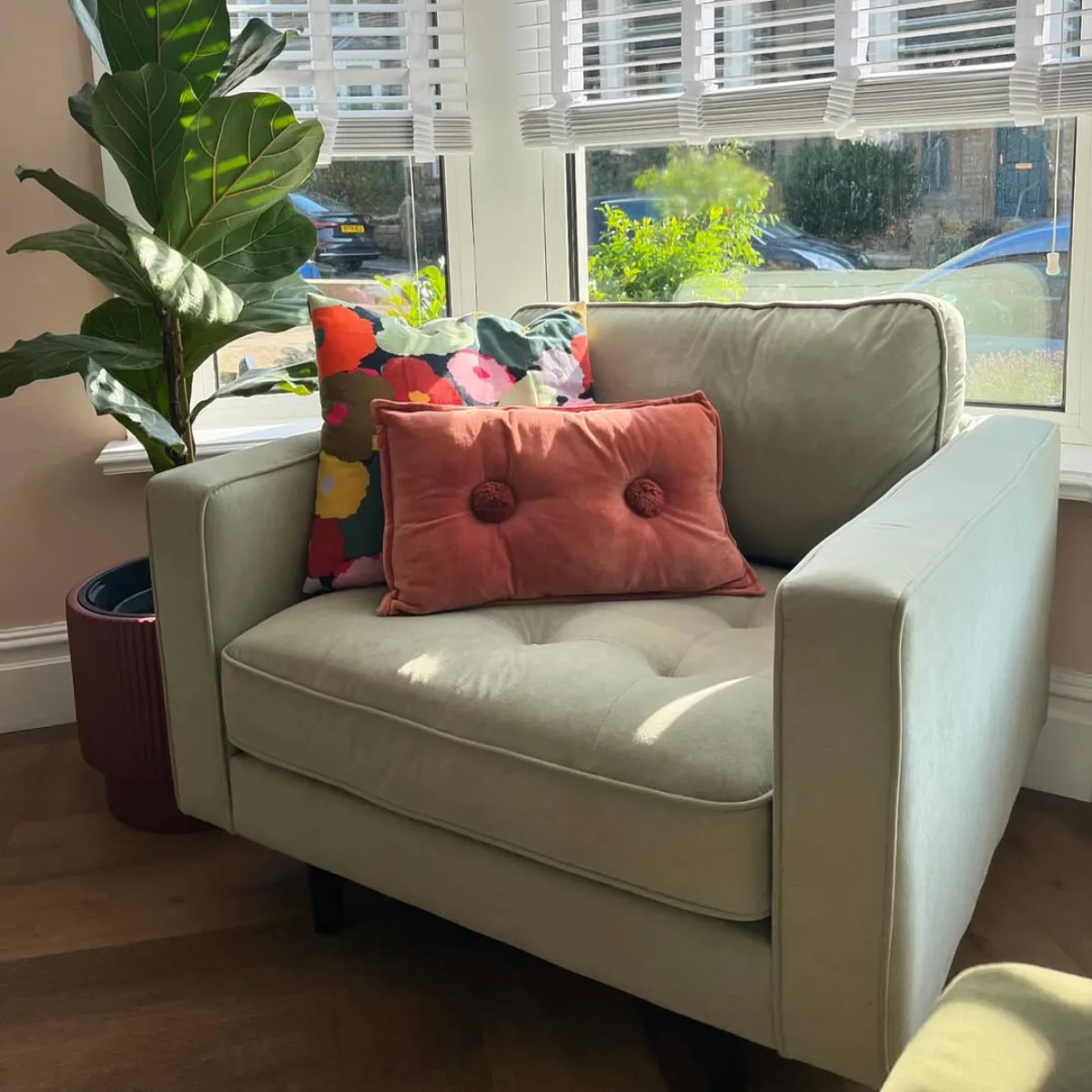 Kursi armchair velvet warna hijau sage dengan bantal dekoratif oranye dan motif bunga di sudut baca dekat jendela yang terang. Cocok untuk bagian artikel yang membahas: Loveseat atau kursi aksen pelengkap.