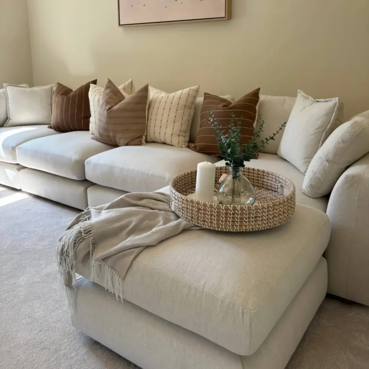 Sofa modular berukuran besar dengan pelapis bahan katun berwarna putih bersih, dihiasi berbagai bantal bernuansa cokelat dan krem, serta nampan rotan berisi lilin dan tanaman di atas sandaran kaki atau ottoman yang empuk.