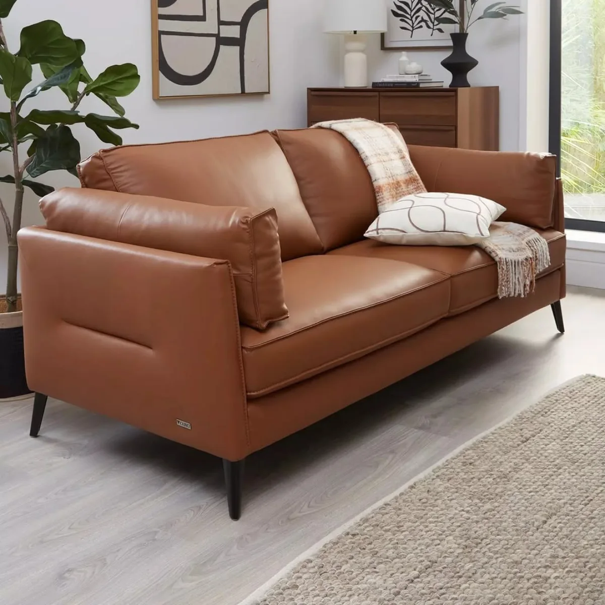 Sofa dua dudukan bergaya modern minimalis yang menggunakan bahan kulit sintetis atau oscar berwarna cokelat halus, ditopang oleh kaki-kaki besi hitam, serta dihiasi selimut hangat dan bantal putih bermotif garis melengkung.