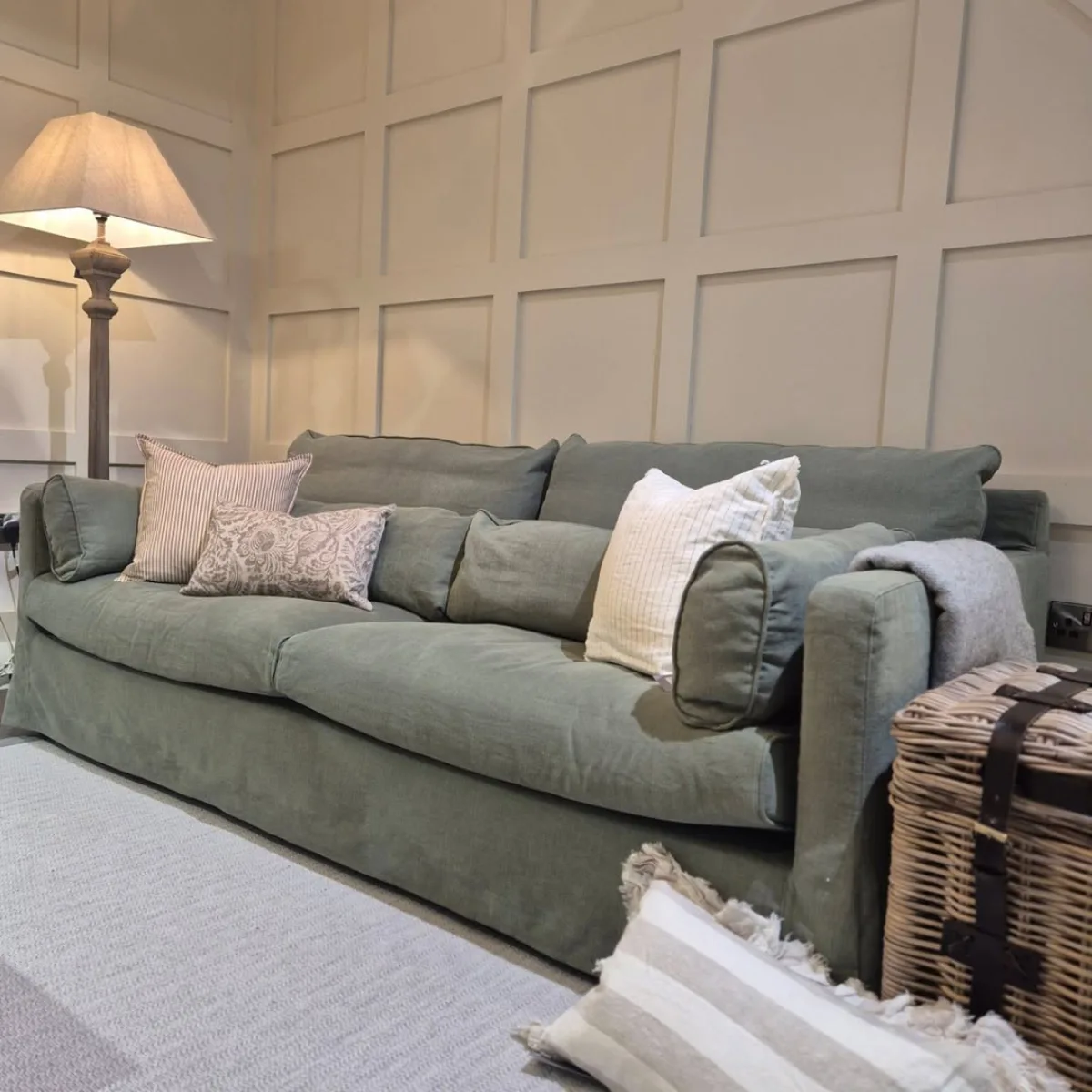 Sofa bergaya santai dengan pelapis bahan linen berwarna hijau sage yang tampak natural dan elegan, dilengkapi dengan beberapa bantal bermotif klasik, diletakkan di depan dinding berpanel kayu dengan lampu lantai di sudut ruangan.