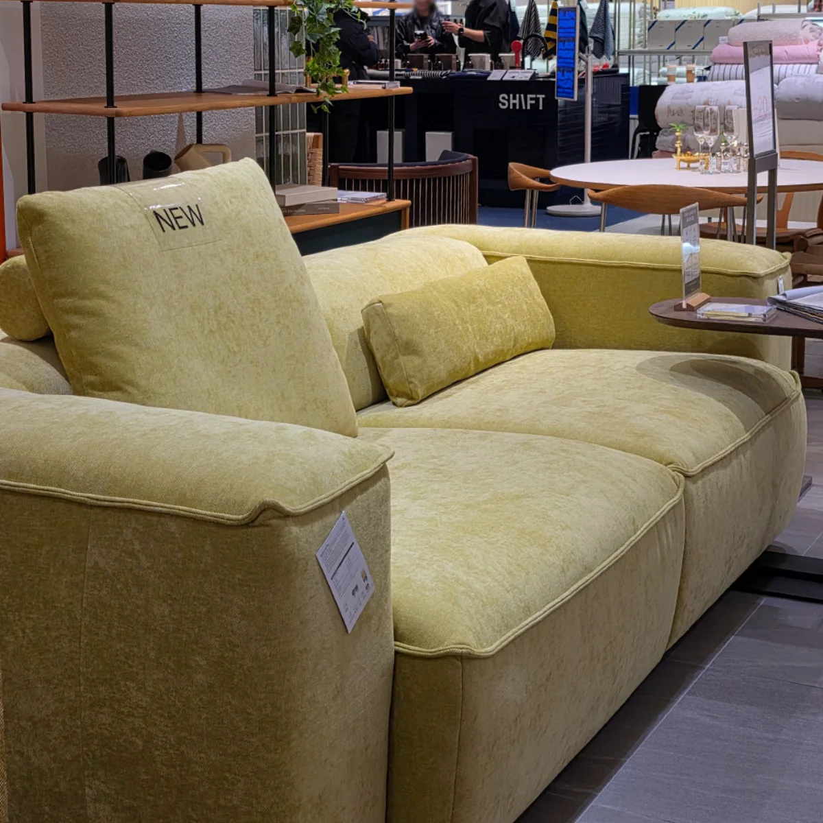 Sofa modern dengan desain tebal dan empuk yang menggunakan bahan polyester berwarna kuning kehijauan cerah, menampilkan tekstur kain yang awet dan kuat, dipajang di area pameran furnitur lengkap dengan bantal sofa senada.