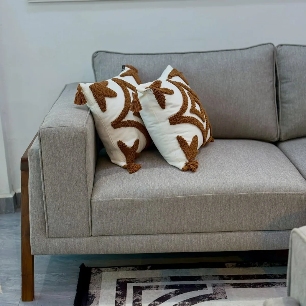 Sofa berdesain kasual dengan pelapis bahan kanvas berwarna abu-abu terang yang kuat dan tahan lama, dikombinasikan dengan bingkai kayu pada bagian lengan, serta dipermanis dengan dua bantal putih berhias motif rumbai cokelat yang estetik.