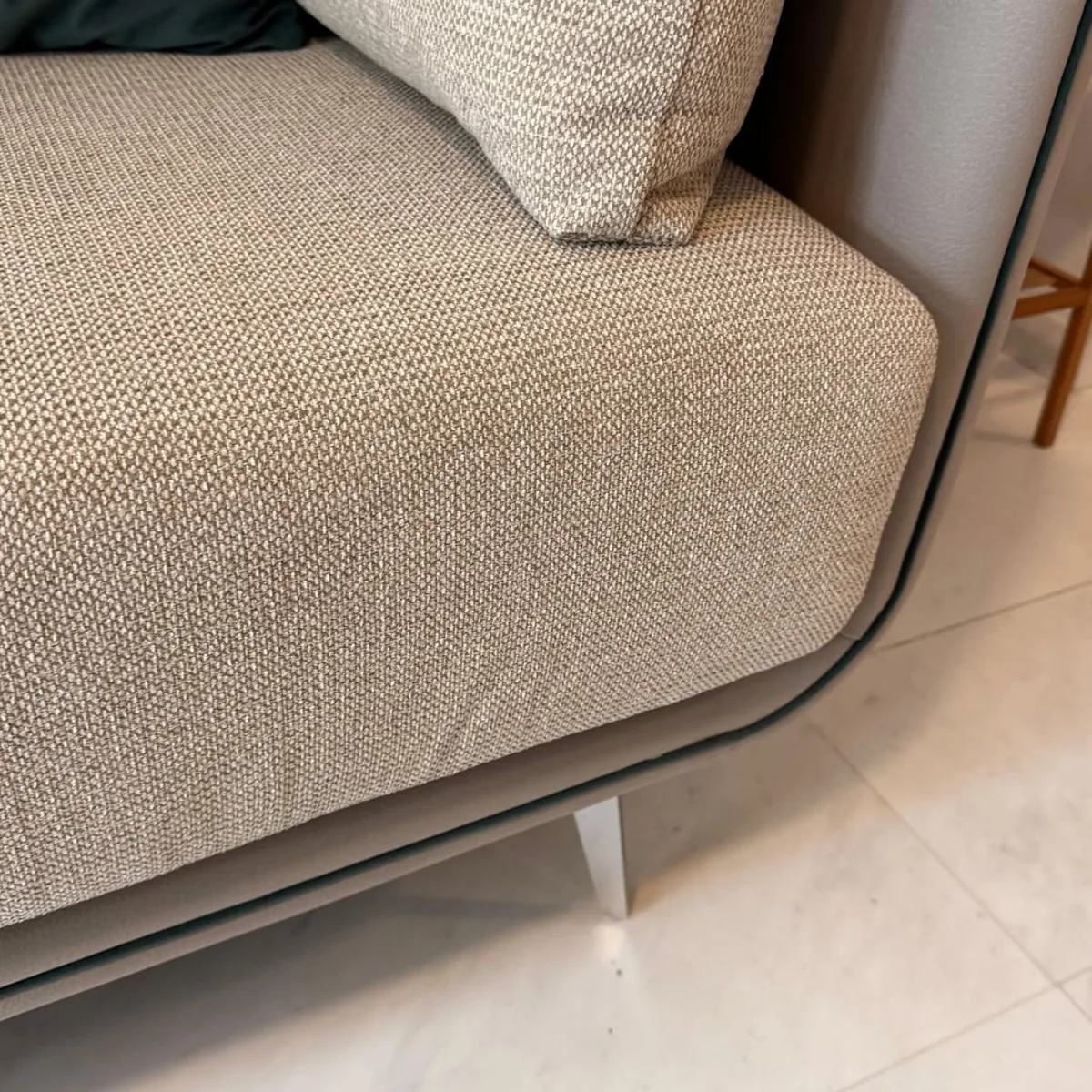 Tampilan jarak dekat dari sudut sofa bersudut melengkung yang menggunakan bahan microfiber berwarna krem keabu-abuan, memperlihatkan tekstur serat kain sintetis yang rapat, halus, dan mudah dibersihkan dari noda.