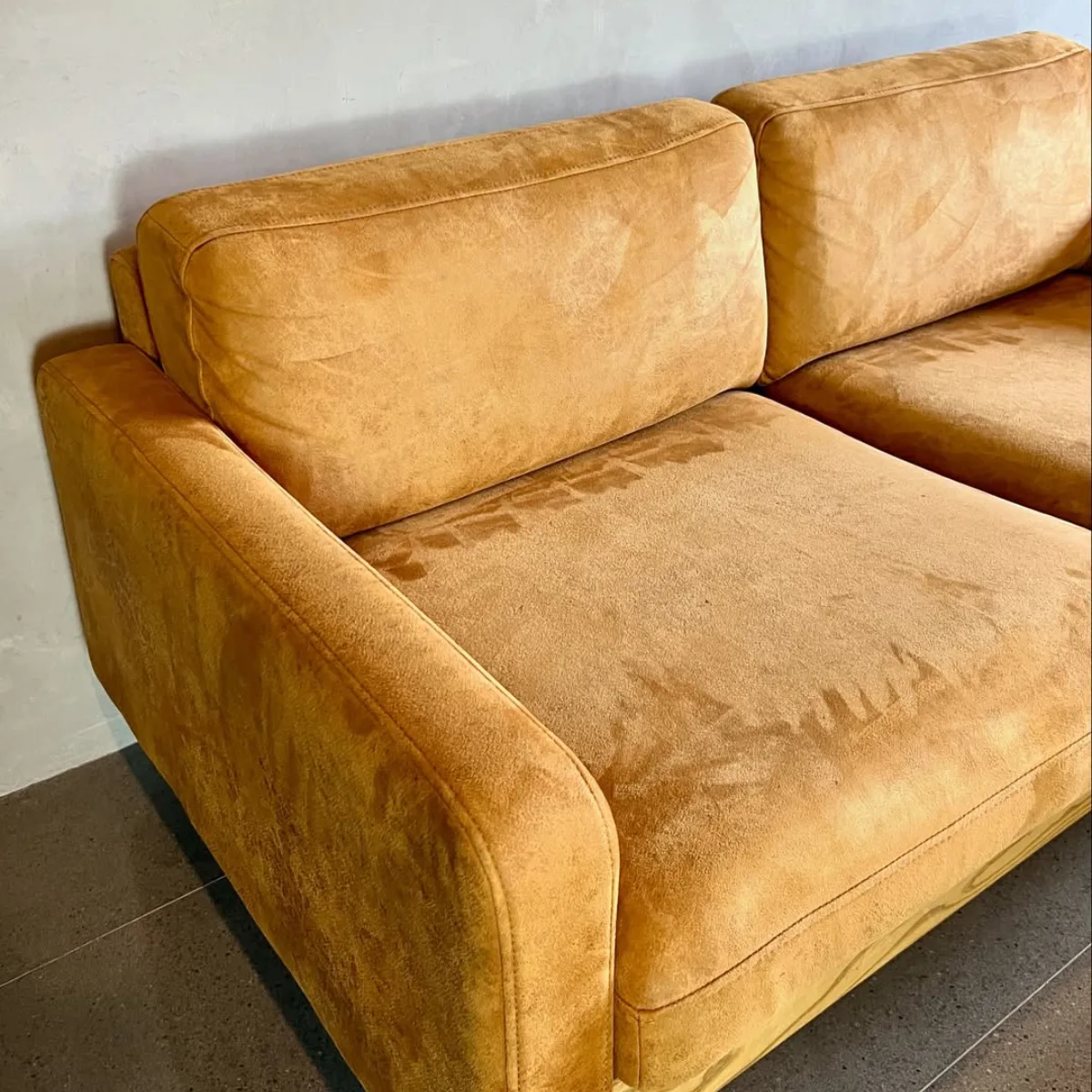 Tampilan detail dari dudukan dan sandaran sofa empuk yang dilapisi bahan suede berwarna kuning mustard yang hangat, memperlihatkan tekstur permukaan matte khas suede yang lembut dan meninggalkan jejak saat diusap tangan.