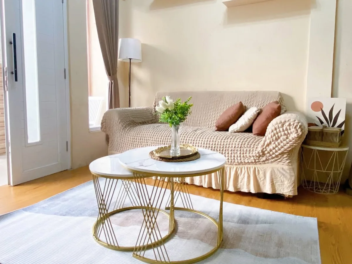 Ruang tamu kecil dengan sofa krem, meja kopi bundar minimalis, karpet netral, dan dekorasi sederhana yang membuat ruangan terlihat rapi dan nyaman.