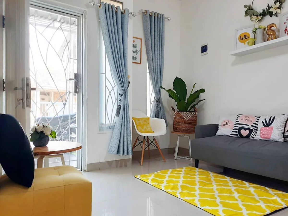 Ruang tamu minimalis dengan sofa abu-abu gelap, karpet kuning cerah, dan gorden biru muda.