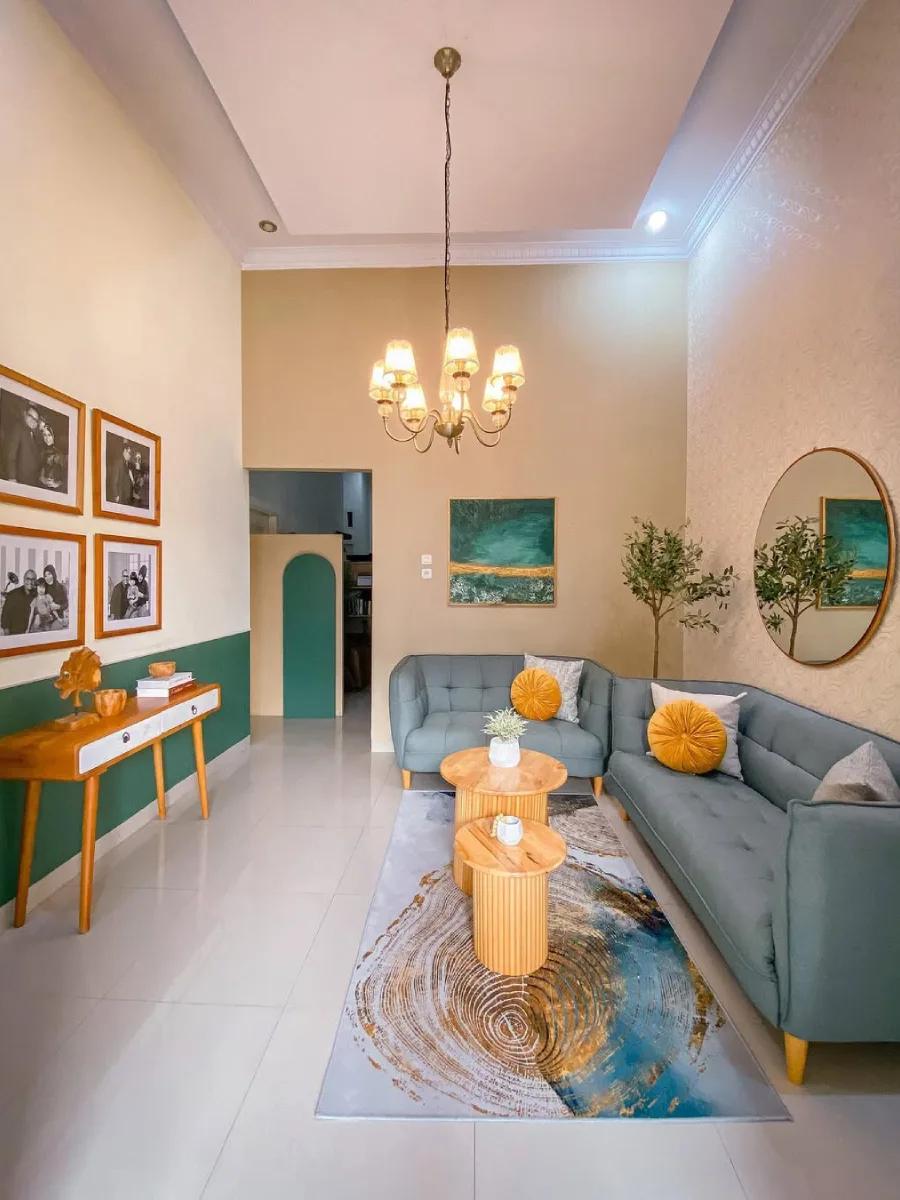 Ruang tamu modern berplafon tinggi (high ceiling) dengan sofa L abu-abu kehijauan, meja kopi kayu bulat bertingkat, cermin bulat besar, dan aksen dinding cat hijau separuh.