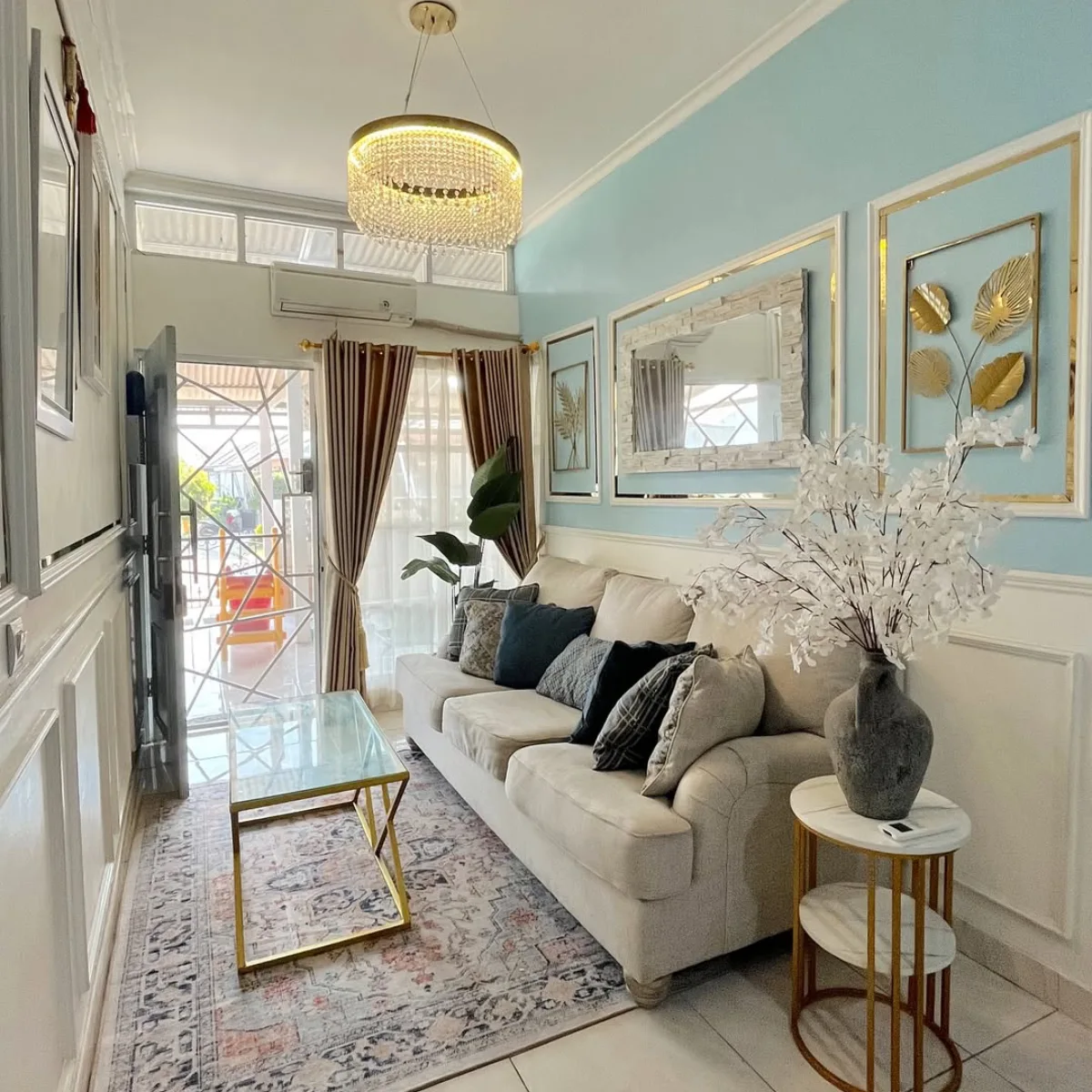 Ruang tamu sempit memanjang nuansa biru pastel dan emas dengan hiasan dinding molding dan lampu gantung kristal.