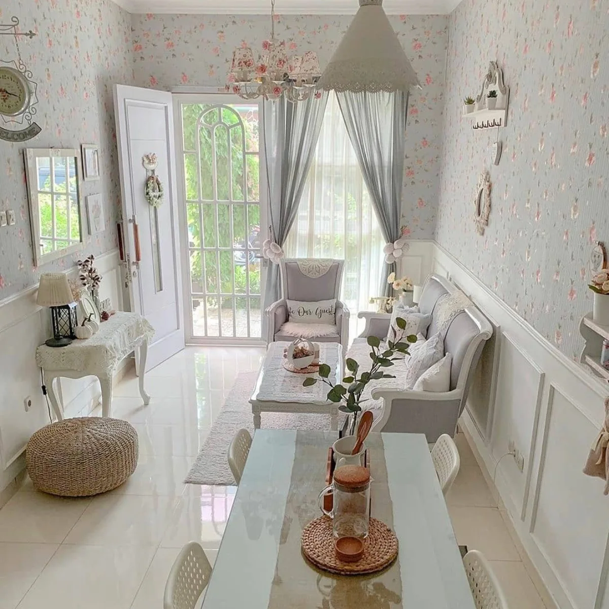 Ruang tamu estetik bergaya shabby chic dengan wallpaper bunga, sofa putih, dan lampu gantung kristal.