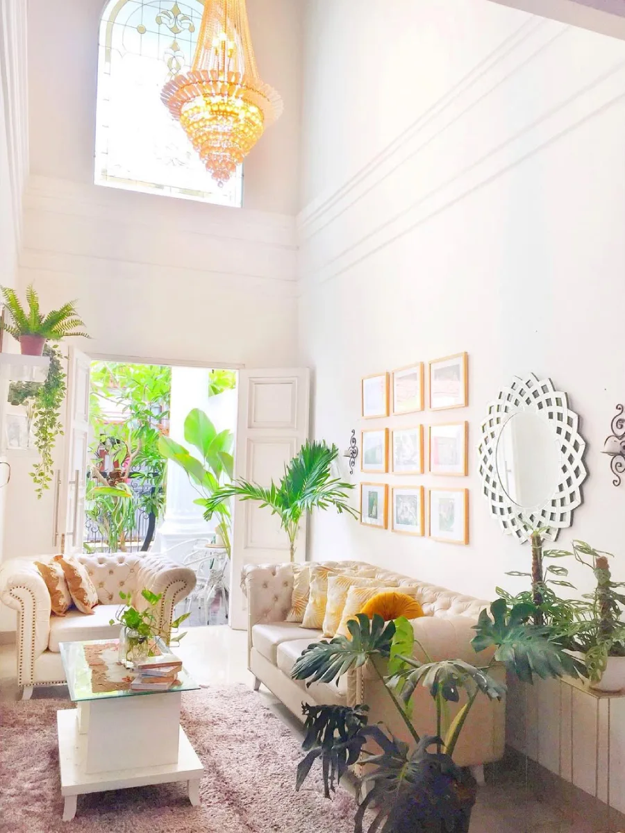 Ruang tamu mewah dengan plafon tinggi (high ceiling), sofa chesterfield klasik, dan tanaman hias indoor yang asri.