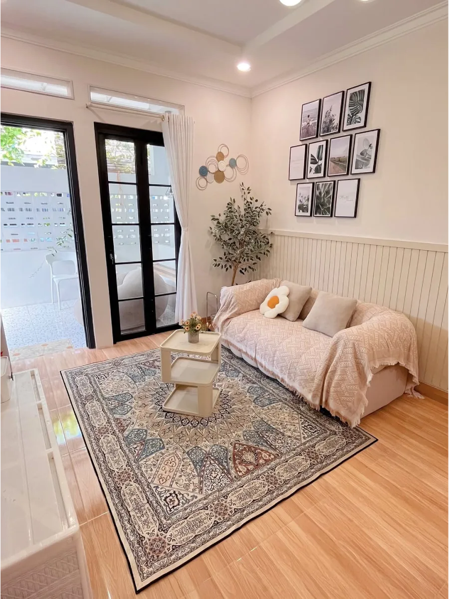 Ruang tamu bergaya scandi-boho dengan karpet bermotif etnik, dekorasi dinding gallery wall bingkai hitam, sofa dengan selimut throw, dan pintu kaca lipat hitam menuju area outdoor.