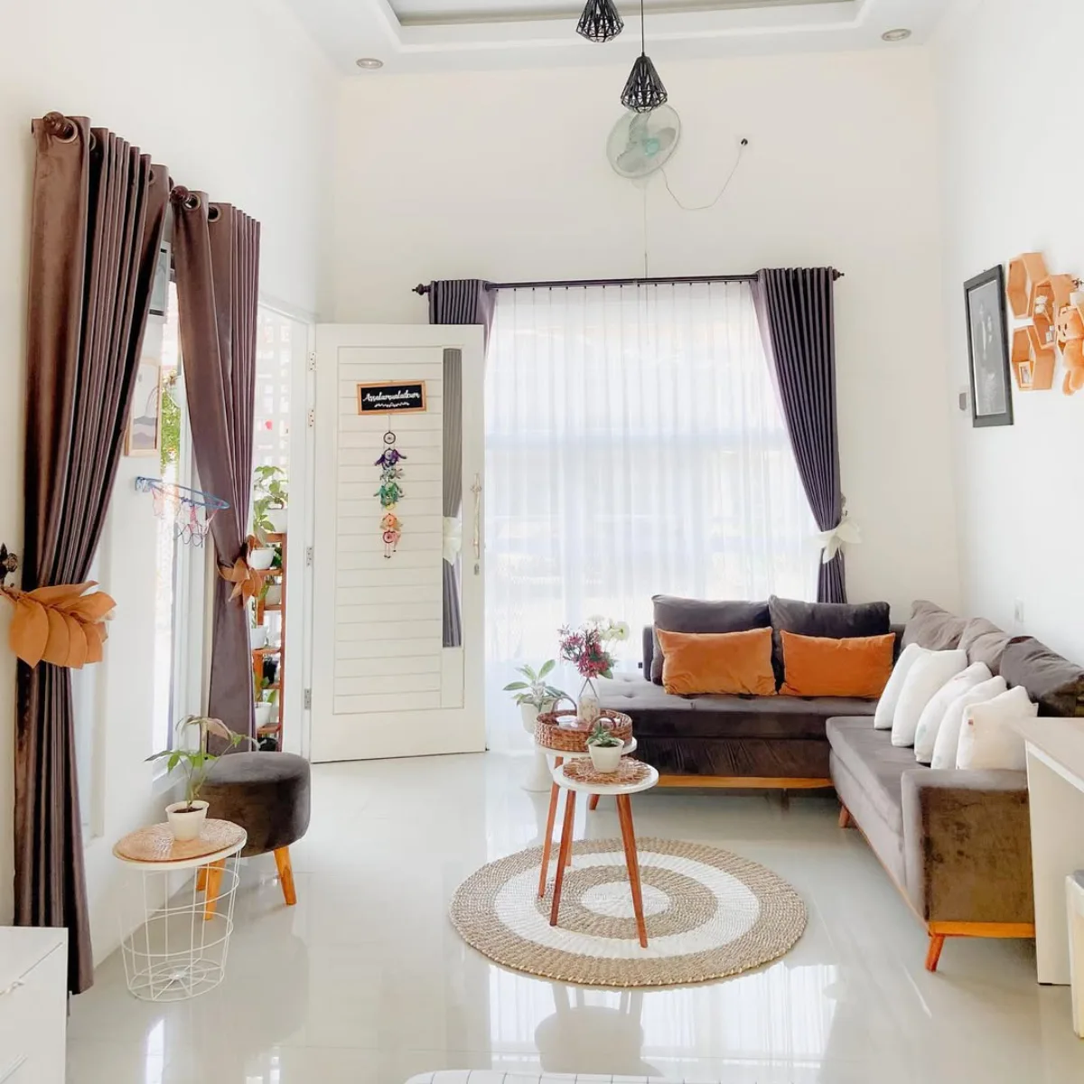 Desain ruang tamu nuansa warna bumi (earth tone) dengan sofa cokelat tua, gorden vitrase putih, dan karpet anyaman bulat.