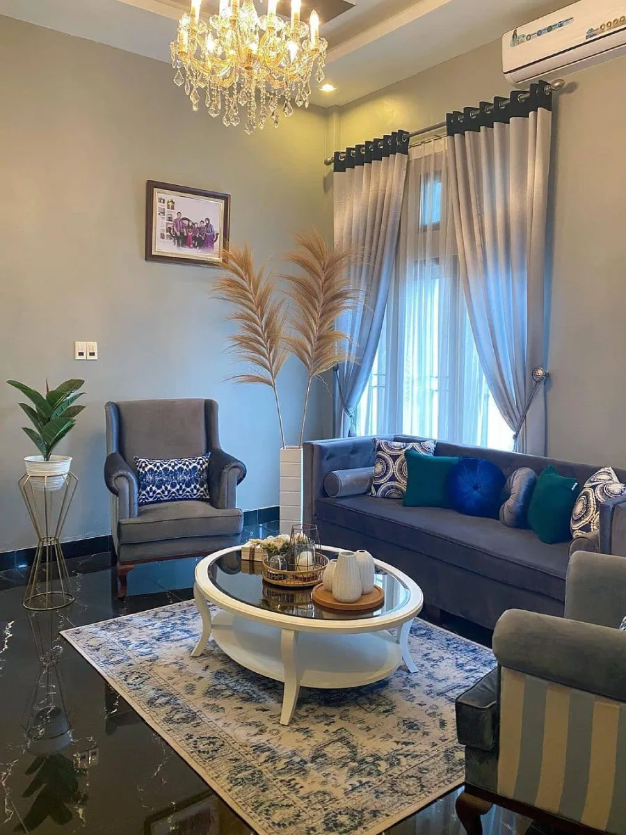 Desain ruang tamu mewah dengan lampu gantung kristal, sofa abu-abu beludru, dan dekorasi pampas kering.