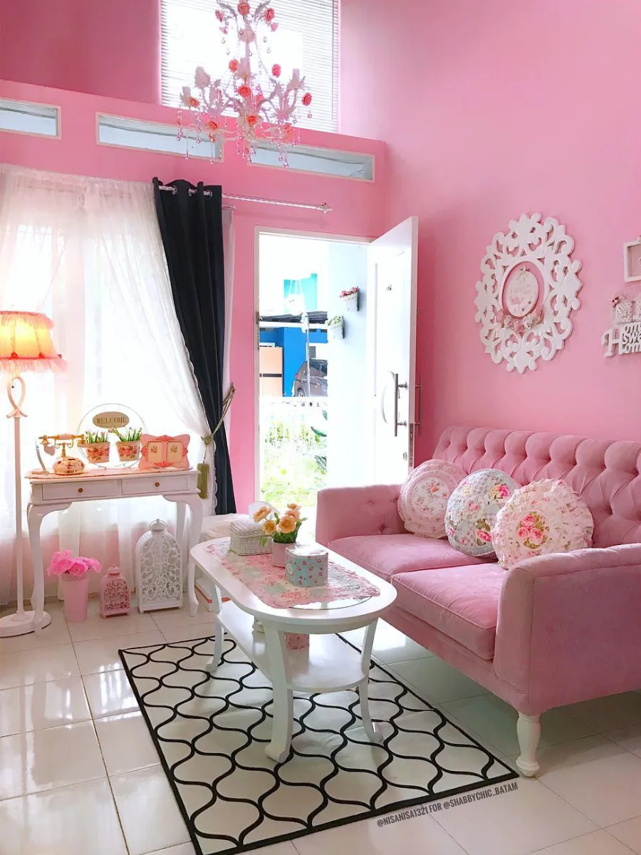 Ruang tamu bernuansa pink shabby chic dengan sofa tufted merah muda, bantal motif bunga, meja konsol putih klasik, dan lampu gantung kristal feminin.
