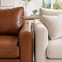 Close-up perbandingan sofa kulit cokelat dan sofa kain beige di ruang tamu modern dengan pencahayaan alami terang.