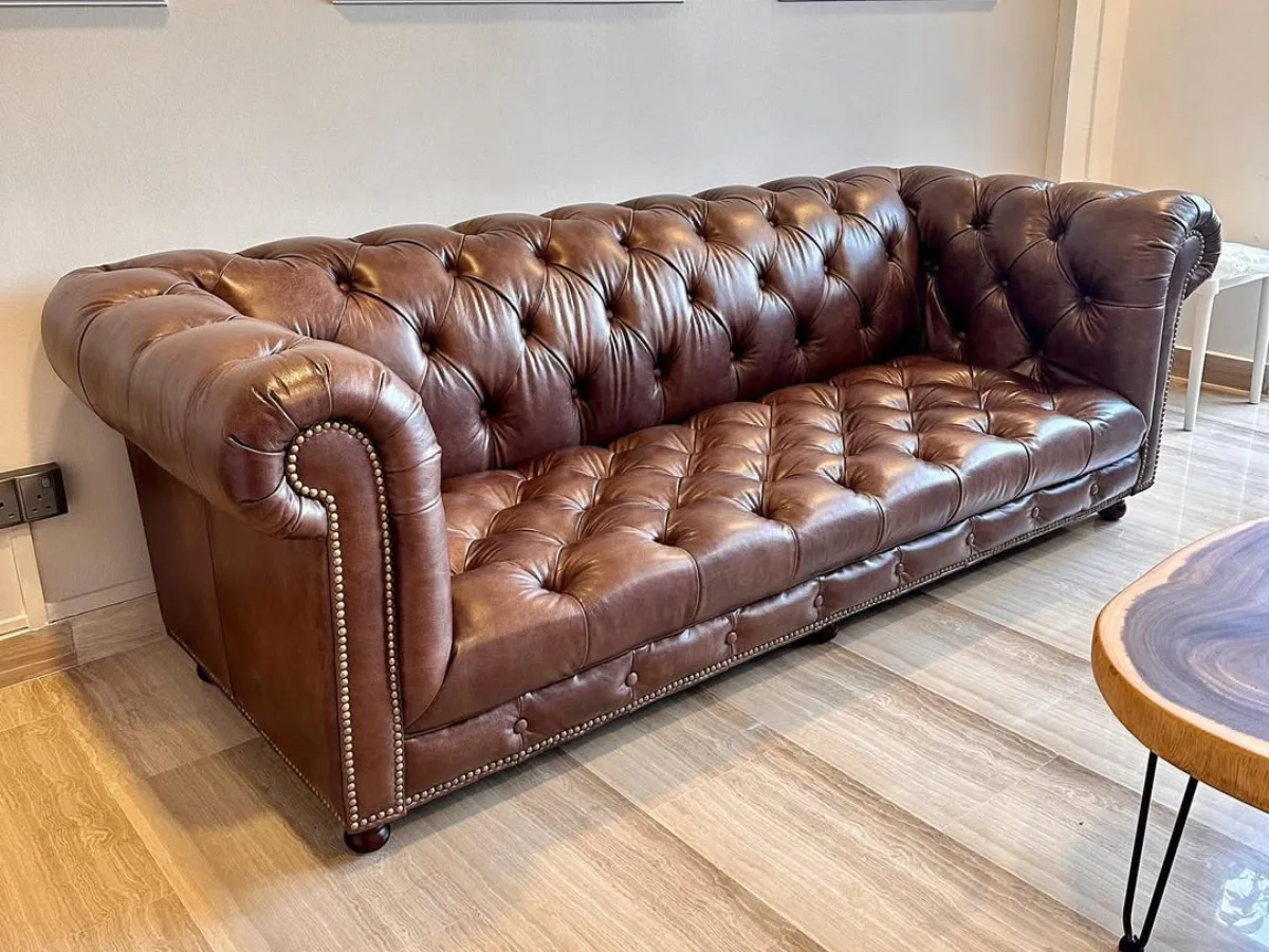 Sofa kulit klasik bergaya Chesterfield berwarna cokelat tua dengan detail kancing dalam (tufting) dan aksen paku payung di bagian lengannya, diletakkan di atas lantai kayu.