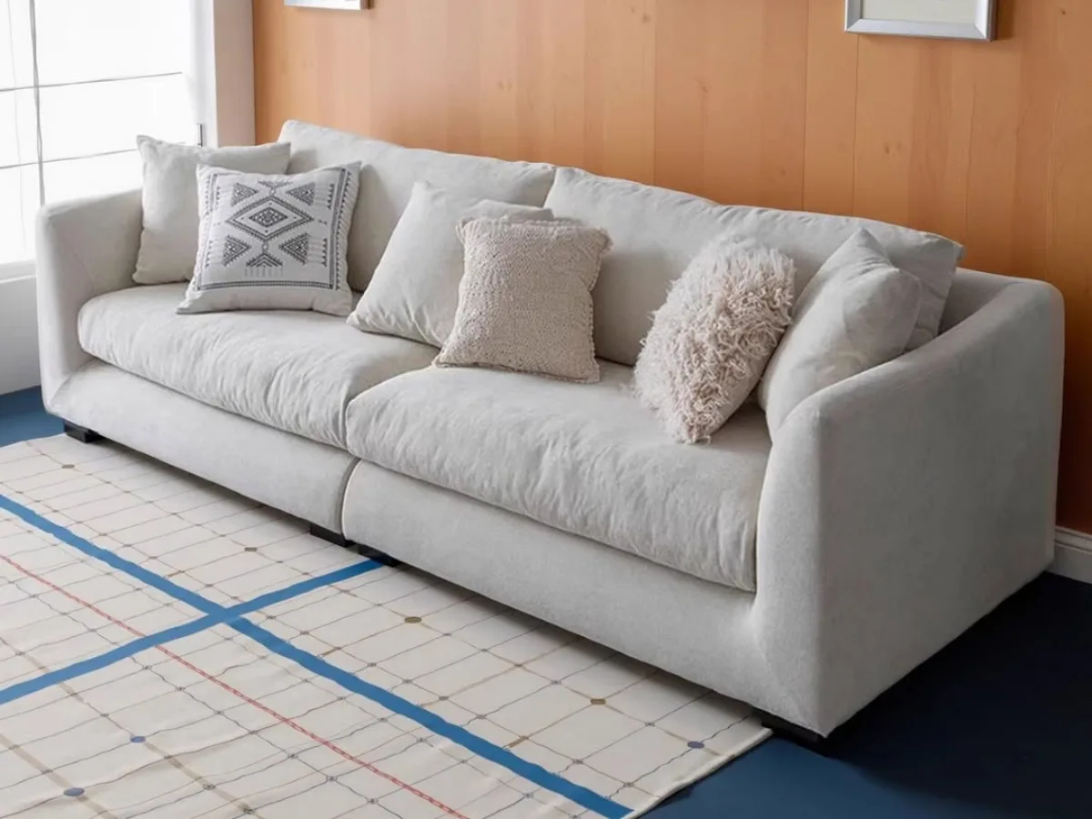 Sofa kain berwarna krem atau putih tulang yang tampak empuk dengan beberapa bantal dekoratif bertekstur dan bermotif, diletakkan di atas karpet berpola garis kotak-kotak.