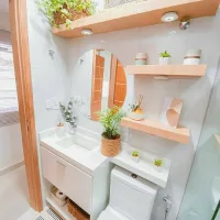 Desain kamar mandi minimalis kecil dengan wastafel menyatu kabinet, toilet duduk, rak dinding kayu, cermin bulat, dan tanaman hias.