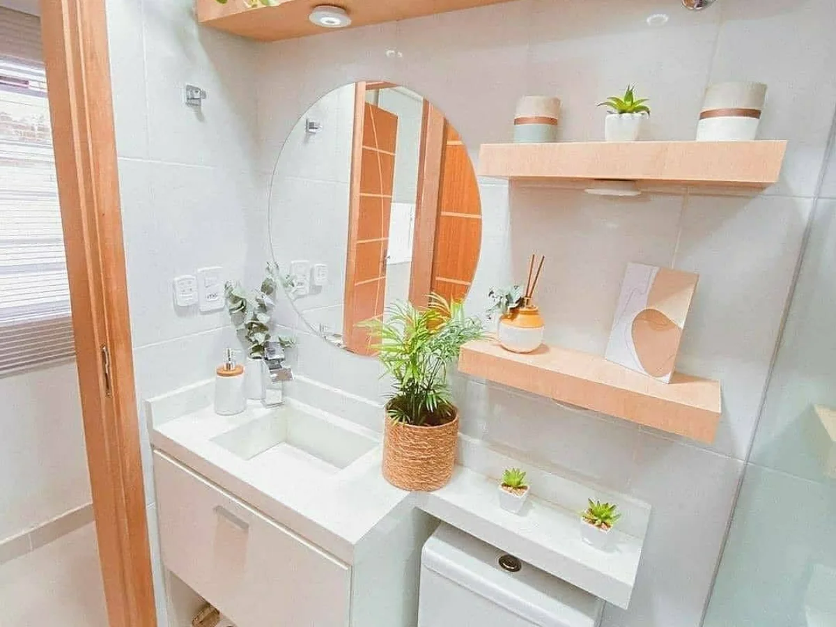 Desain kamar mandi minimalis kecil dengan wastafel menyatu kabinet, toilet duduk, rak dinding kayu, cermin bulat, dan tanaman hias.