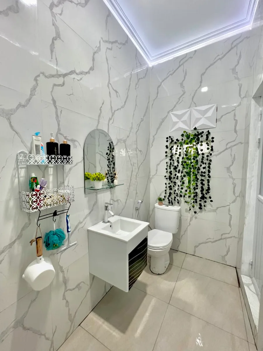 Interior kamar mandi dengan desain minimalis dan modern yang didominasi oleh keramik motif marmer putih cerah. Gambar ini menampilkan area wastafel gantung, cermin oval, rak perlengkapan mandi berwarna putih, serta kloset duduk dengan dekorasi tanaman rambat artifisial di dinding. Penataan ruang ini sangat efisien untuk ukuran  meter, memberikan kesan bersih dan rapi.