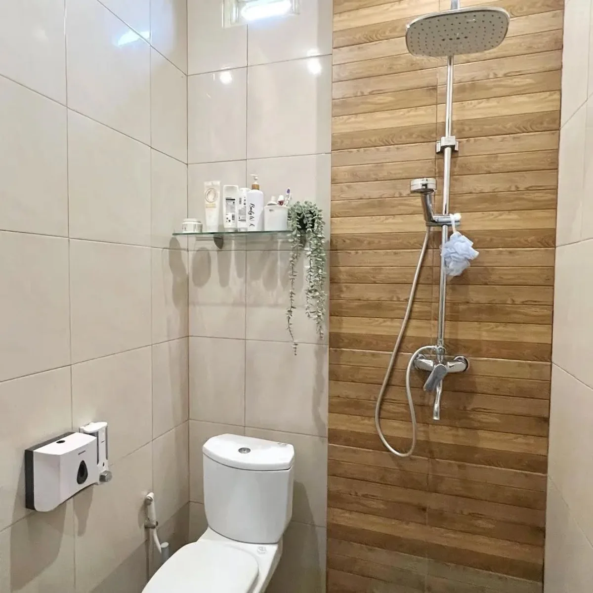 Inspirasi desain kamar mandi minimalis modern ukuran 1.5 x 1.5 meter yang dilengkapi dengan area shower beraksen dinding motif kayu, toilet duduk putih, dan rak kaca untuk perlengkapan mandi agar terlihat rapi.