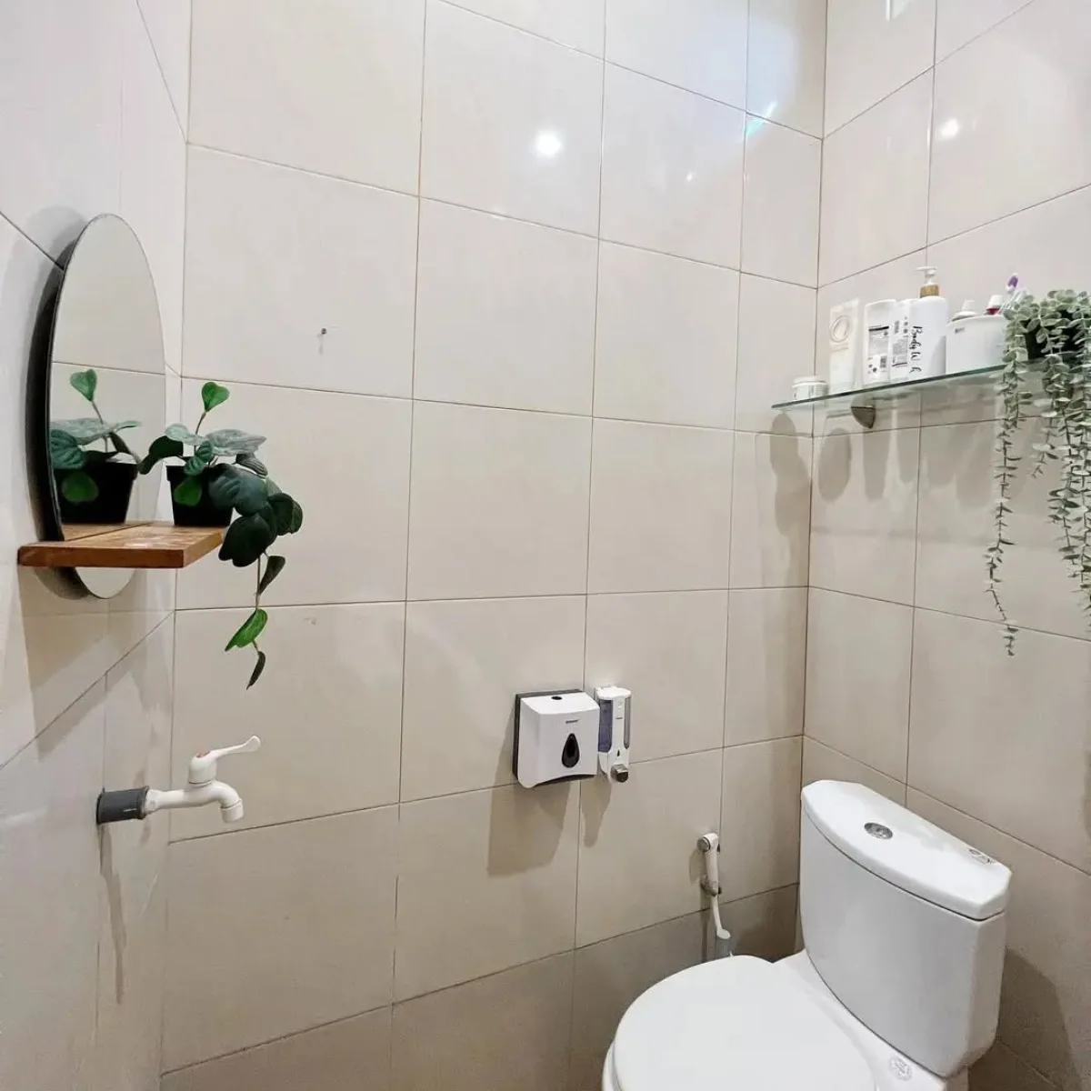 Interior desain kamar mandi minimalis modern ukuran 1.5 x 1.5 meter dengan dinding keramik krem, cermin bulat dengan tanaman hias, serta tata letak ruang yang efisien dan nyaman dipandang.