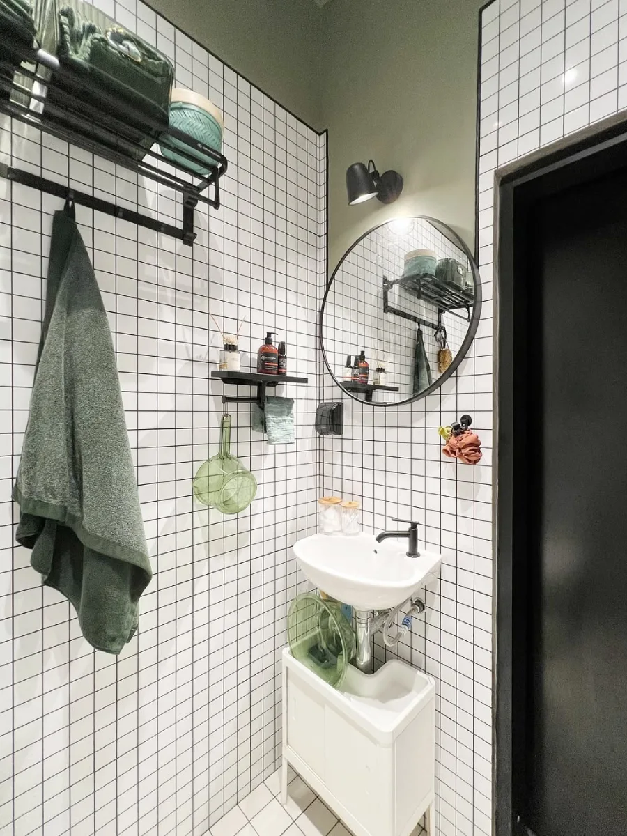Inspirasi desain kamar mandi minimalis modern berukuran 1.5 x 1.3 meter dengan wastafel gantung putih, cermin bulat hitam, dan dinding keramik motif kotak-kotak kecil yang rapi.