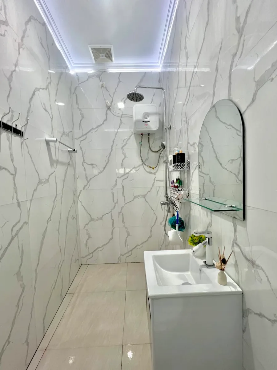 Sudut pandang area shower pada kamar mandi bergaya modern dan minimalis. Terlihat instalasi water heater listrik, rain shower stainless steel, dan gantungan kapstok hitam pada dinding keramik marmer putih. Tata letak perabotan sanitasi ini dirancang khusus untuk memaksimalkan fungsi ruang pada ukuran  meter agar tetap terasa lega.