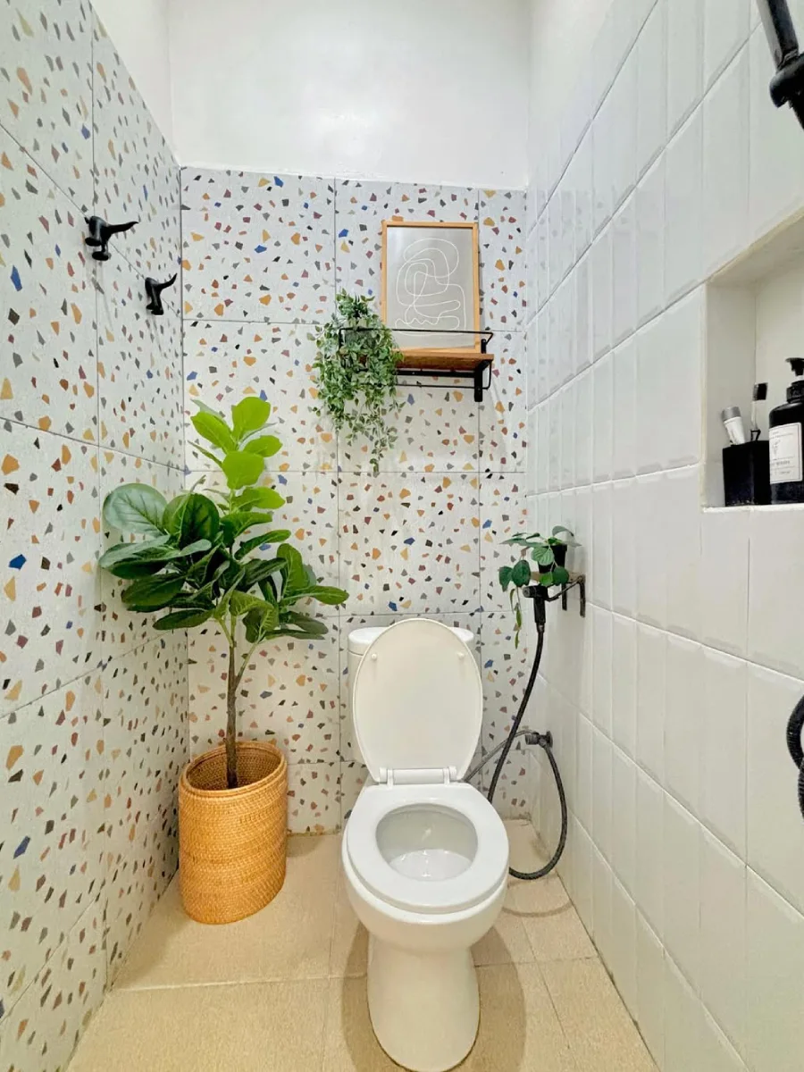 Foto sudut pandang luas desain kamar mandi minimalis modern berukuran 1.6 x 1.2 meter yang menampilkan dinding keramik motif terrazzo warna-warni, kloset duduk putih, serta dekorasi tanaman hijau dalam pot rotan yang memberikan kesan segar dan rapi.