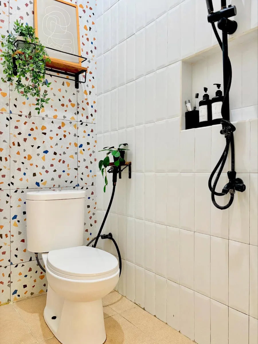 Foto close-up desain kamar mandi minimalis modern ukuran 1.6 x 1.2 meter yang memperlihatkan detail dinding ubin putih bersih dengan rak dinding niche untuk perlengkapan mandi, serta paduan dinding aksen terrazzo yang estetik di area kloset.