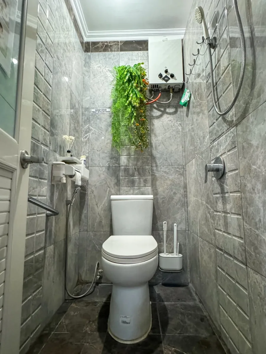 Foto desain kamar mandi minimalis modern berukuran 1 x 1.6 meter dengan dinding keramik motif marmer abu-abu. Ruangan terlihat rapi dengan wastafel compact, kloset duduk putih, tanaman hias gantung yang memberi kesan segar, serta pencahayaan yang terang.