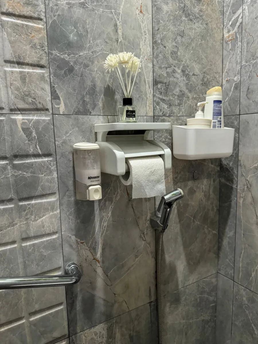 Detail aksesoris pada desain kamar mandi minimalis modern ukuran 1 x 1.6 meter, menampilkan rak dinding putih yang efisien. Di atasnya terdapat dekorasi bunga kering, pengharum ruangan, dispenser sabun cair, dan tempat tisu toilet yang tersusun rapi untuk memaksimalkan ruang terbatas.