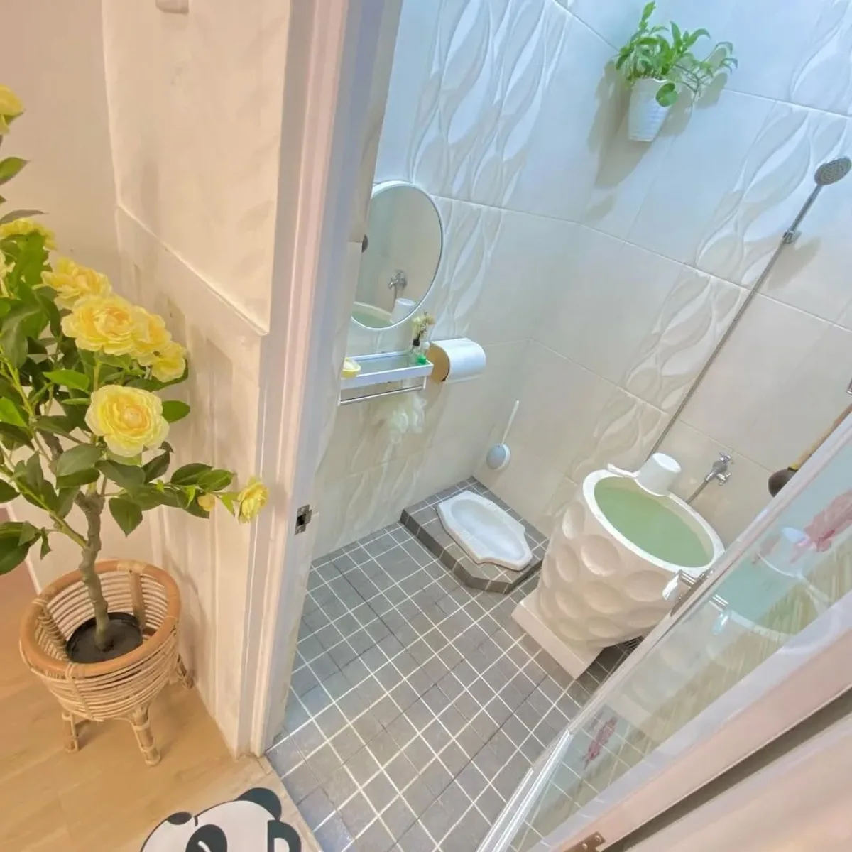 Inspirasi desain kamar mandi minimalis modern ukuran 1.2 x 1.5 meter dengan kloset jongkok, bak mandi teraso unik, dan dekorasi bunga kuning yang memberikan kesan estetik dan rapi.