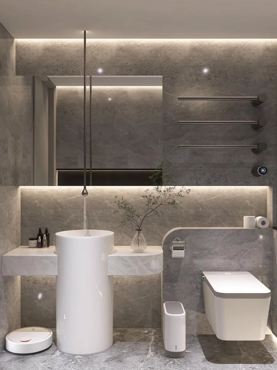 Sudut pandang area wastafel pada kamar mandi bernuansa abu-abu modern. Terdapat wastafel berbentuk silinder putih yang berdiri bebas (freestanding) dengan kran air yang turun dari langit-langit. Cermin besar berbentuk persegi panjang dipasang di dinding dengan lampu latar (backlight) hangat, serta gantungan handuk minimalis berwarna hitam di sisi kanan.