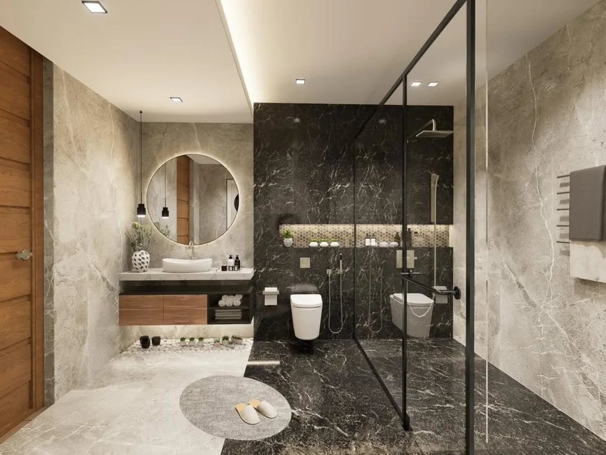 Interior kamar mandi kontemporer yang memisahkan area basah dan kering. Area shower menggunakan marmer hitam pekat, dibatasi oleh kaca bening berbingkai hitam. Area kering menggunakan ubin abu-abu terang dengan meja wastafel kayu gantung, cermin bulat dengan lampu LED, dan hiasan batu koral putih di bawah kabinet.