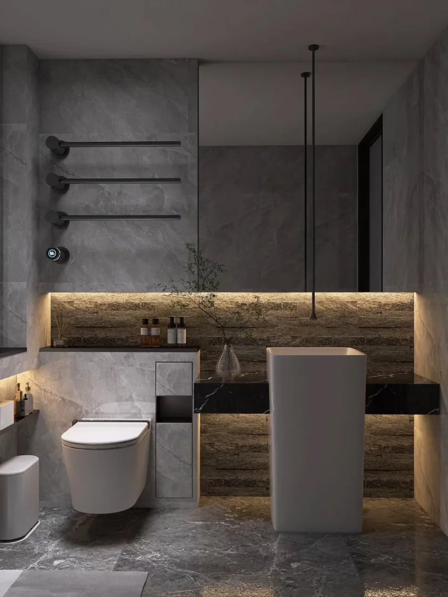 Area wastafel kamar mandi minimalis dengan wastafel pedestal (berdiri) berbentuk kotak putih, cermin besar tanpa bingkai, toilet gantung dinding, dan rak handuk hitam matte yang terpasang di dinding batu abu-abu bertekstur.