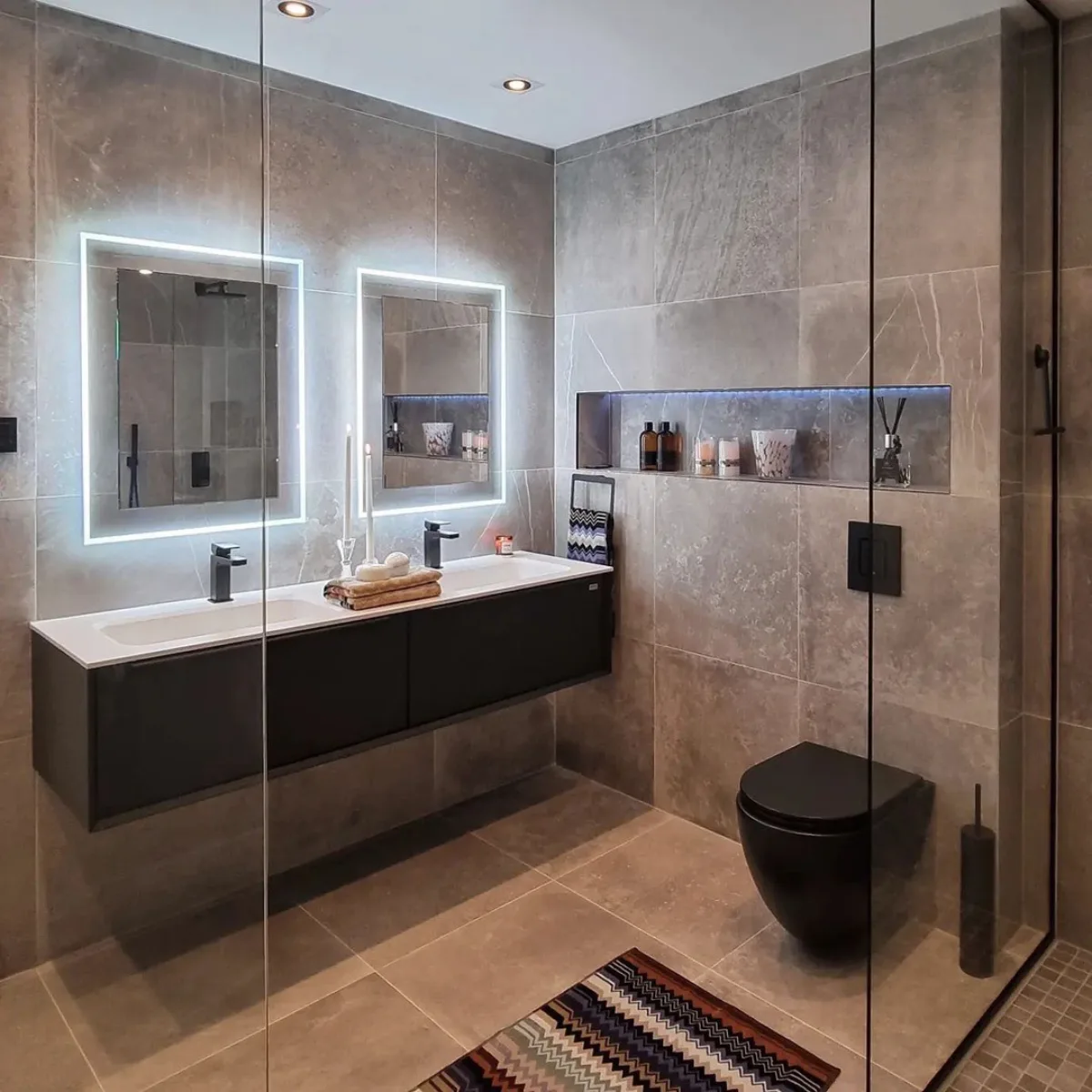 Sudut pandang menyamping dari area wastafel ganda. Terlihat jelas dua cermin dengan bingkai cahaya putih terang. Di dinding belakang toilet hitam, terdapat ceruk (niche) panjang berlapis marmer abu-abu dengan lampu biru muda untuk menaruh dekorasi dan pewangi ruangan.