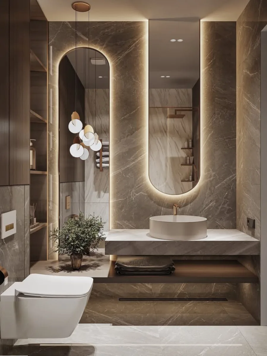 Desain wastafel kamar mandi mewah bernuansa hangat dengan dinding marmer cokelat, cermin berbentuk lengkung (arch) dengan lampu latar LED, wastafel bulat di atas meja batu, dan lampu gantung kluster modern yang elegan.