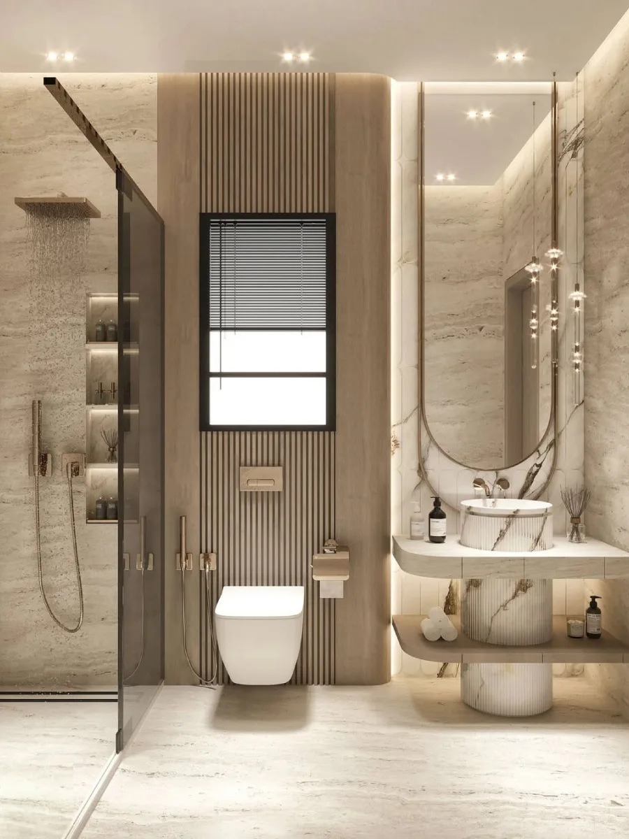 Kamar mandi bernuansa spa alami dengan dinding batu travertine beige, wastafel pedestal marmer bertekstur alur (fluted), cermin oval panjang, dan dinding aksen kayu vertikal di belakang toilet gantung.