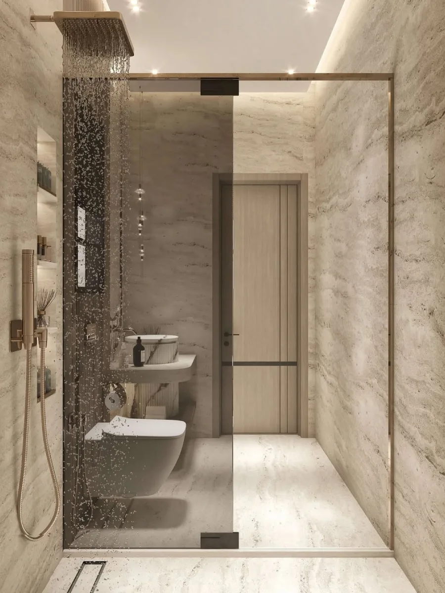 Sudut pandang dari dalam area shower yang memperlihatkan dinding batu alam beige, kepala shower hujan yang tertanam di plafon, pintu kaca geser minimalis, dan pencahayaan hangat yang menenangkan.