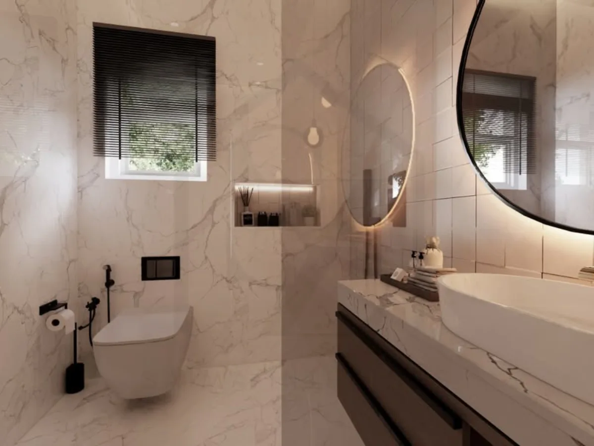 Close-up area wastafel kamar mandi marmer putih, memperlihatkan detail keran dinding, ceruk (niche) dinding untuk sabun dengan pencahayaan LED, dan pantulan cahaya alami dari jendela di cermin bulat.