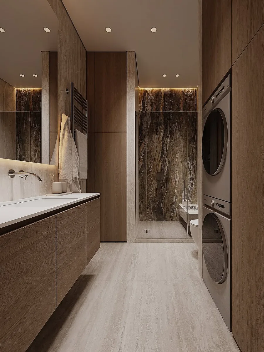 Interior kamar mandi mewah dengan dominasi panel kayu hangat, dilengkapi mesin cuci dan pengering bertumpuk (laundry area), serta meja wastafel panjang dengan pencahayaan lembut.
