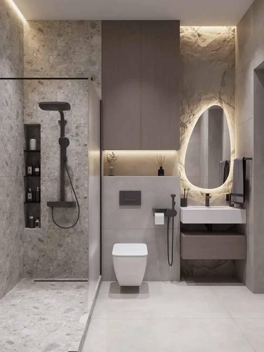 Kamar mandi bergaya minimalis industrial dengan ubin teraso abu-abu, cermin bentuk organik (asimetris) dengan lampu belakang, dan rak dinding tanam vertikal.