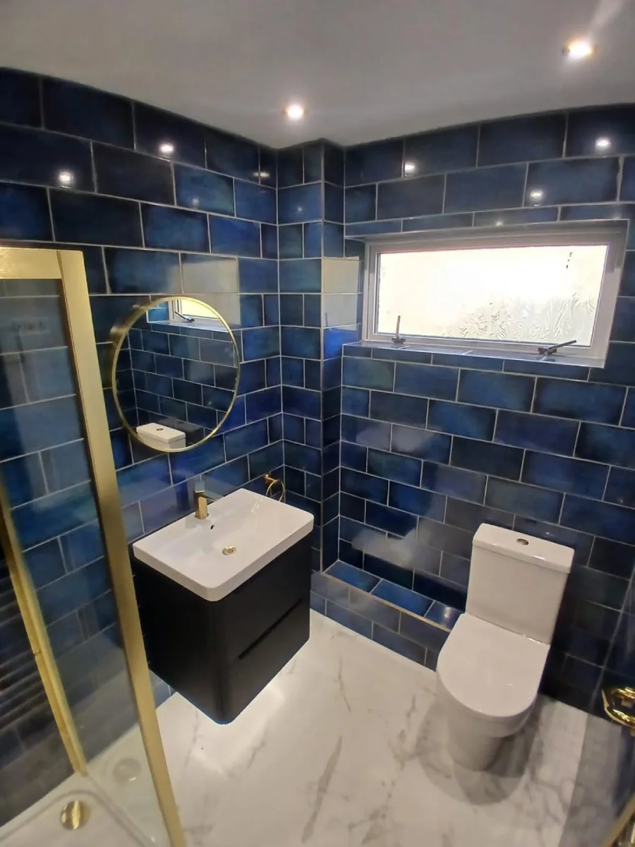 Kamar mandi kecil dengan ubin dinding model bata (subway tiles) berwarna biru tua mengkilap, lantai marmer putih, wastafel kabinet hitam modern, cermin bulat berbingkai emas, dan jendela kecil di bagian atas untuk ventilasi.