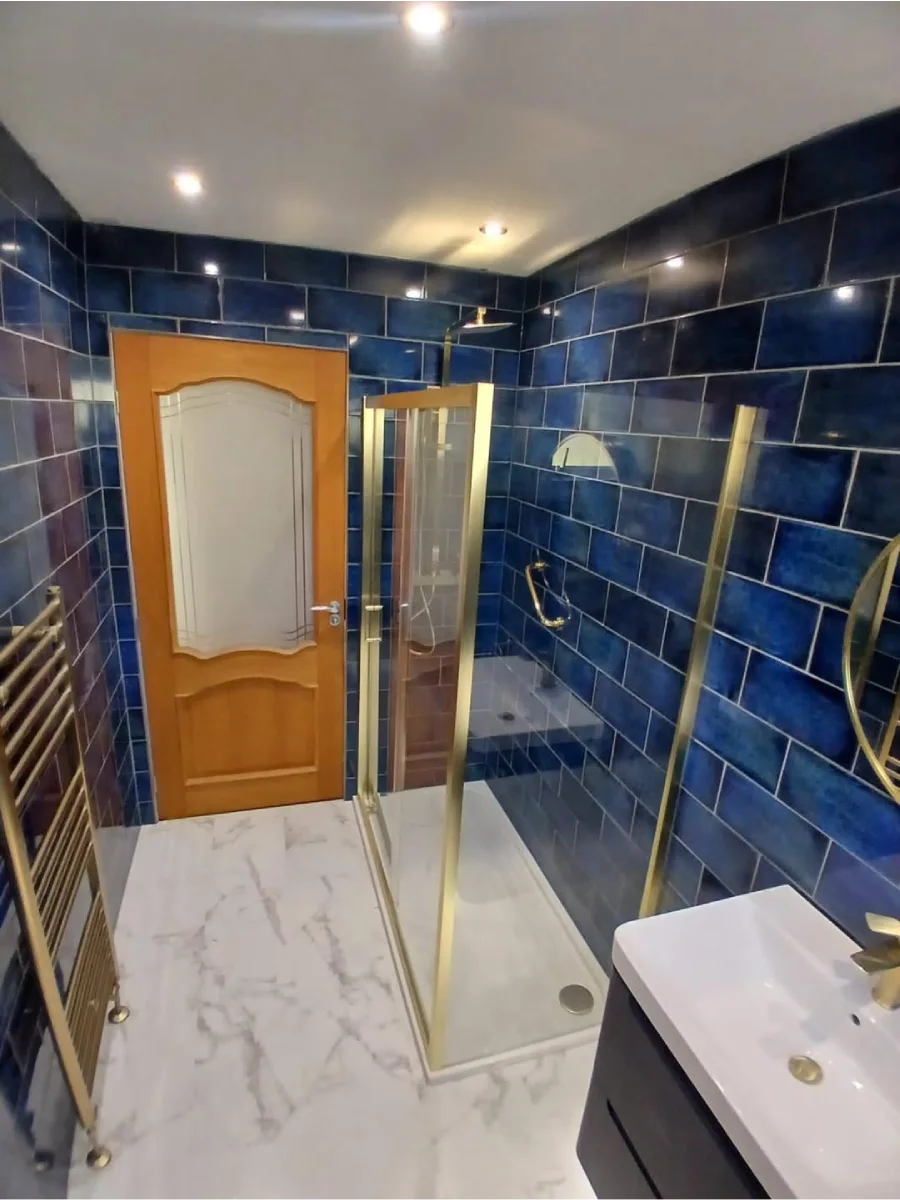 Interior kamar mandi dengan bilik shower kaca berbingkai emas, gantungan handuk pemanas (heated towel rail) berwarna emas, pintu kayu klasik dengan kaca buram, yang kontras dengan dinding keramik biru tua yang dominan.